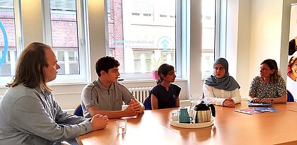 Fünf Menschen sitzen um einen Tisch in einer Interviewsituation. Es steht Cafe und Tee auf der Tisch.